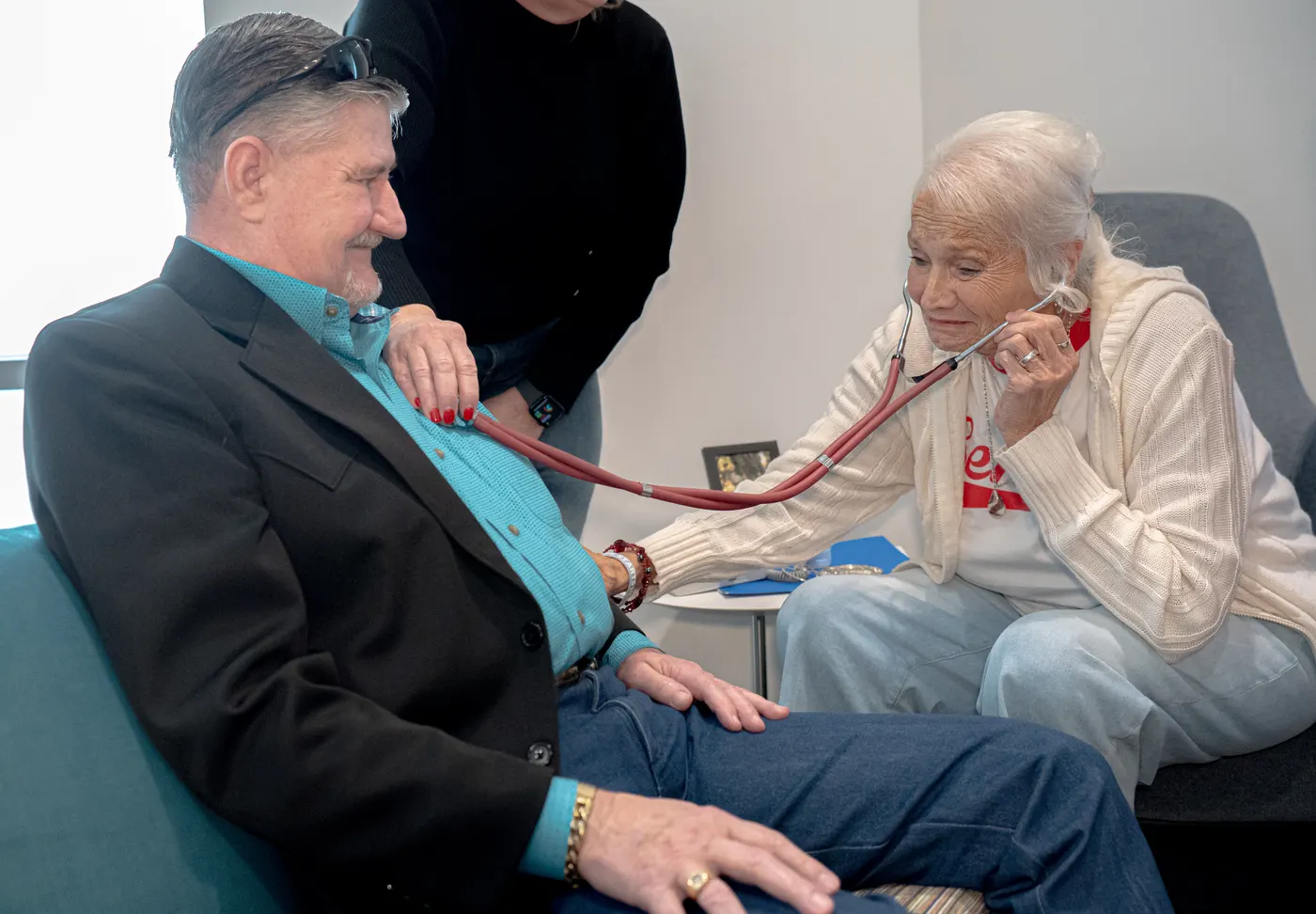 An older woman listens through a stethoscope placed on a man’s chest as he sits beside her.
